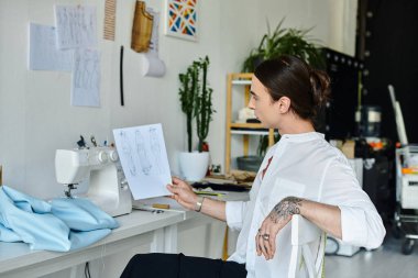 Atölyesinde çalışan genç bir adam, aletler ve kumaşlarla çevriliyken bir kıyafet tasarımını inceliyor ve sürdürülebilir modaya olan bağlılığını gösteriyor..