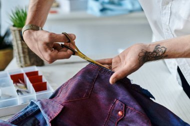 Genç bir adam, atölyesinde altın makasla bir kumaş parçasını dikkatlice kesiyor ve eski kıyafetleri yeniden canlandırma sanatını vurguluyor..