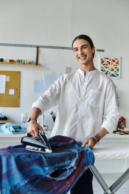 Genç bir tasarımcı, giysi restorasyon atölyesindeki kot gömleğini ütülerken gülümsüyor ve eski eşyalara yeni bir hayat veriyor..