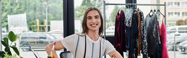 A young gay man smiles confidently in his clothing restoration atelier, surrounded by garments awaiting transformation.