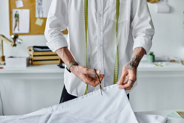 A young man in a white shirt uses scissors to cut fabric while wearing a tape measure around his neck. He is in a workshop and works as a clothing restoration specialist.