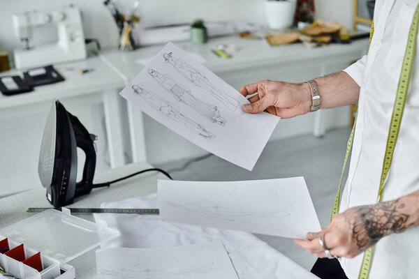A young gay man, dressed in a white shirt, reviews clothing sketches in his DIY clothing restoration atelier.