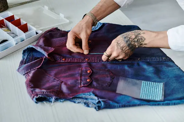 Stock image A young man with tattoos works on a discarded denim shirt, giving it new life with sustainable clothing restoration.