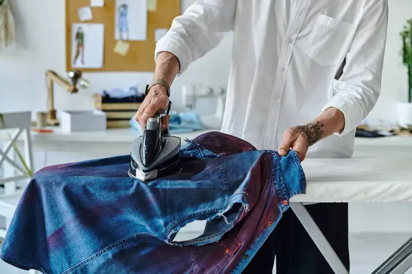 stock image A young man in a white shirt carefully irons a denim shirt in his atelier.