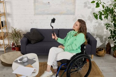 Tekerlekli sandalyedeki genç bir esmer kadın oturma odasında selfie çubuğuyla video çekerken gülümsüyor..