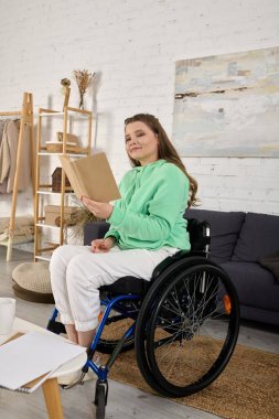 Kahverengi saçlı genç bir kadın oturma odasında tekerlekli sandalyede oturmuş kitap okuyor..