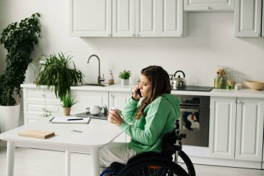 Tekerlekli sandalyedeki genç esmer kadın mutfaktaki bir masada oturuyor, elinde bir kupayla telefonuyla konuşuyor..