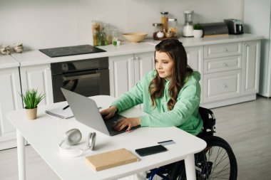 Tekerlekli sandalyedeki esmer bir kadın oturma odasında oturmuş dizüstü bilgisayarla çalışıyor..