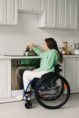 Kahverengi saçlı genç bir kadın mutfaktaki tekerlekli sandalyede oturur ve ocakta çaydanlığa uzanır..