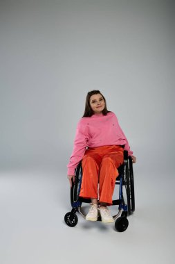 Siyah saçlı genç bir kadın stüdyo ortamında tekerlekli sandalyede oturuyor, pembe bir kazak ve turuncu pantolon giyiyor..