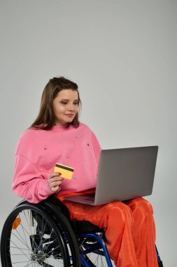 A young woman in a wheelchair sits in a studio setting, using a laptop and holding a credit card. clipart