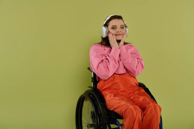 Tekerlekli sandalyedeki genç esmer bir kadın stüdyo ortamında kulaklıkla müzik dinliyor. Pembe bir kazak ve turuncu pantolon giyiyor..