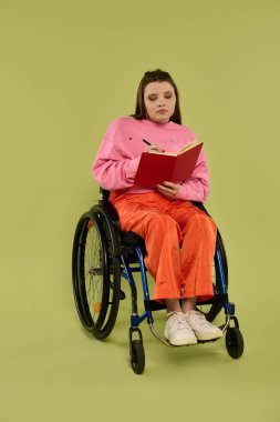Tekerlekli sandalyedeki genç bir kadın düşünceli bir şekilde stüdyoda kırmızı bir deftere yazıyor..