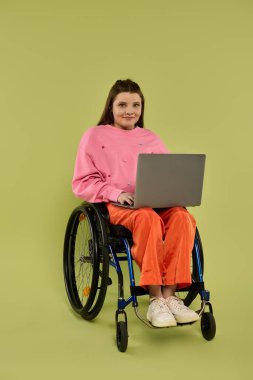 A young brunette woman in casual attire sits in a wheelchair, using a laptop in a studio setting with a green backdrop. clipart