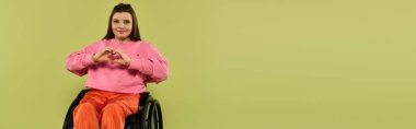 A young woman with brown hair sits in a wheelchair in a studio setting, smiling and making a heart shape with her hands. clipart