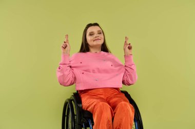 A young brunette woman in a wheelchair sits in a studio with a green background. She is wearing casual attire and has her fingers crossed. clipart