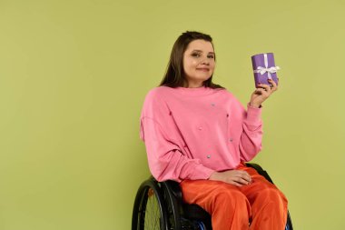 Kahverengi saçlı genç bir kadın stüdyo ortamında tekerlekli sandalyede oturuyor. Pembe bir kazak ve turuncu pantolon giyiyor ve elinde beyaz bir fiyonklu mor bir hediye kutusu var..