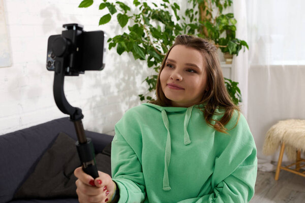 A young woman sits in her living room, smiling as she films a video with her phone on a selfie stick.