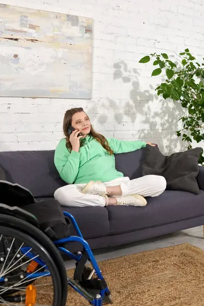 stock image A young woman in a wheelchair sits on a couch in her living room, talking on her phone.