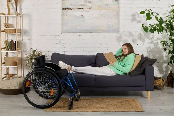 stock image A young brunette woman with a disability relaxes on a sofa in her living room, reading a book. Her wheelchair sits beside her.