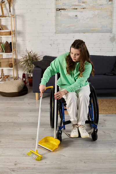 A young woman in a wheelchair sweeps the floor of her living room with a broom and dustpan.