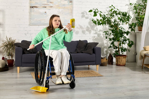 A young brunette woman sits in a wheelchair in her living room, holding a broom and sweeping the floor.