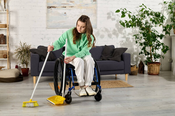 A young woman in a wheelchair is sweeping the floor of her living room. She is smiling and looking down at the broom.
