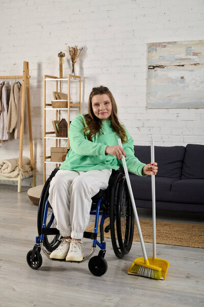 A young woman in a wheelchair is smiling as she holds a broom in her living room.