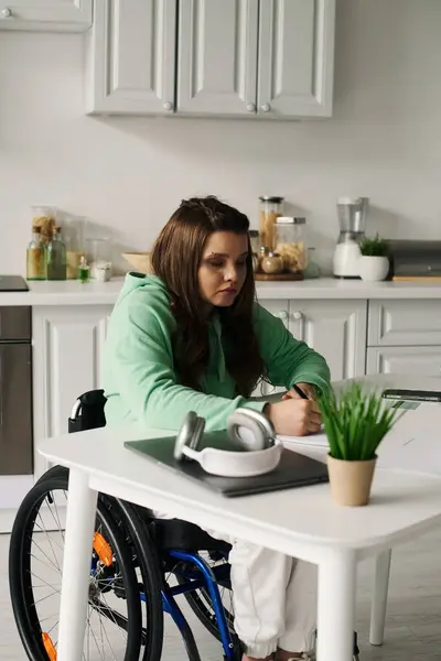 stock image A young woman in a wheelchair is working at a table in her home. She is wearing a green sweatshirt and is writing on a piece of paper.