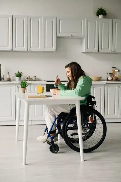 stock image A young brunette woman with a disability sits in a wheelchair and eats breakfast at a white table in her home.