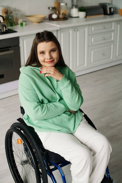A young woman with long brown hair sits in a wheelchair in her kitchen, her relaxed posture and calm expression suggest a moment of quiet reflection.