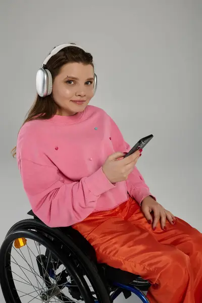 stock image A young brunette woman with a disability sits in a wheelchair while wearing headphones and holding a phone in a studio setting.