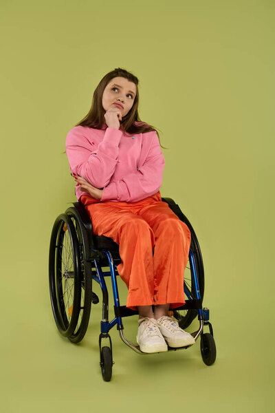 A young woman with long brunette hair sits in a wheelchair in a studio setting. She wears casual clothing and appears to be deep in thought.