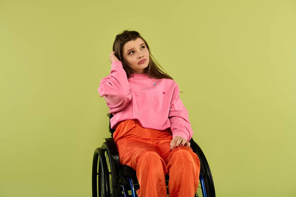 A contemplative brunette young woman with a disability, seated in a wheelchair, rests her head in her hand in a studio setting.
