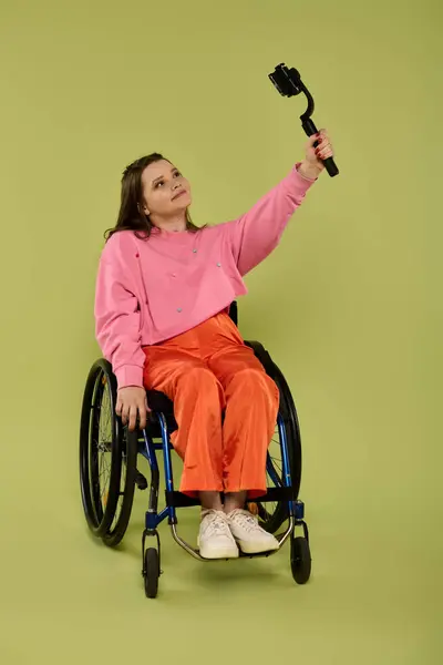 stock image A young woman in a wheelchair smiles and takes a selfie with a selfie stick in a studio setting.