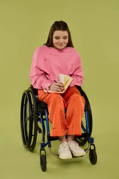 stock image A young woman with long brunette hair sits in a wheelchair in a studio setting, writing in a notebook.