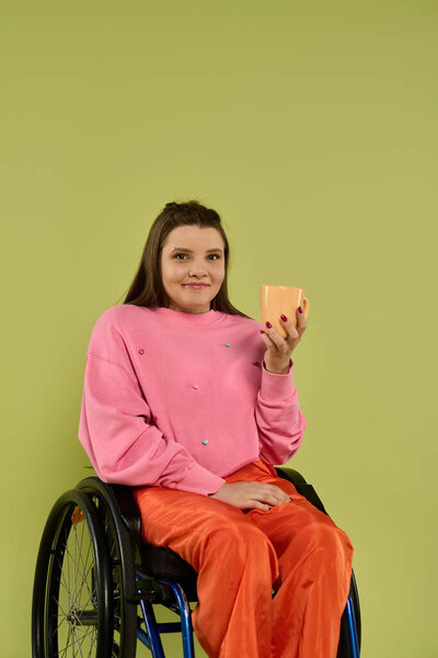 A young brunette woman in casual clothing sits in a wheelchair against a green backdrop, holding a light orange mug in her right hand.