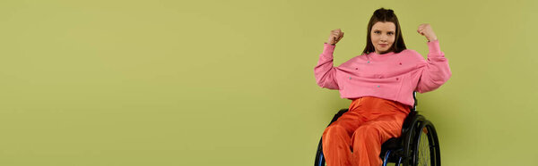 Молодая женщина с длинными каштановыми волосами, одетая в розовый свитер и оранжевые штаны, садится в инвалидное кресло и сгибает бицепс в студии.