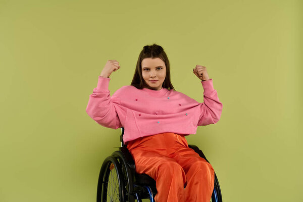 Молодая брюнетка с инвалидностью сидит в инвалидном кресле в студии. Она одета в повседневную одежду и позирует с поднятыми руками, уверенно.