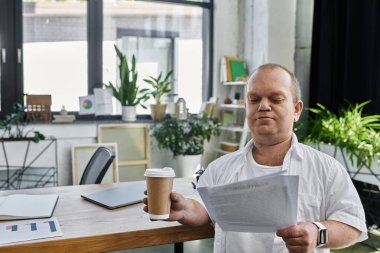 Elinde bir fincan kahve olan bir adam modern ofis ortamında dokümanları gözden geçirir..