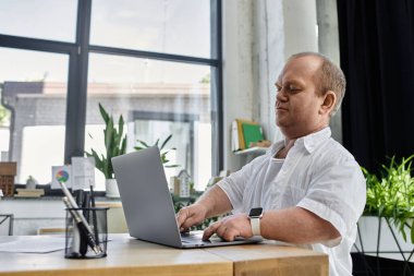 Beyaz gömlekli bir adam masada oturur ve ofis ortamında dizüstü bilgisayar kullanır..