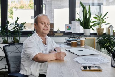Kapsamlı bir adam, modern bir ofiste masa başında işine odaklanmış olarak oturur. Arka plandaki doğal ışık ve yeşillik sakin ve üretken bir atmosfer yaratır..
