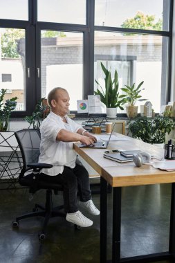 Kapsamlı bir adam, modern bir ofiste, büyük pencereli bir masada oturuyor ve bir dizüstü bilgisayar üzerinde çalışıyor..