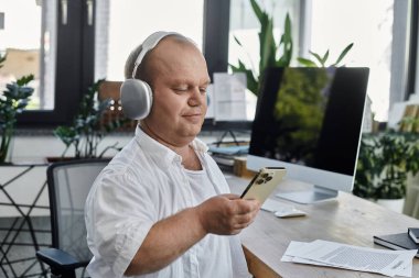 Kapsayıcı kulaklık takan bir adam ofisinde, masasında çalışıyor ve akıllı telefonunu kullanıyor..