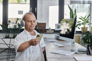 Kapsayıcı kulaklık takan bir adam ofisinde masasında oturmuş, akıllı telefonunu kullanıyor..
