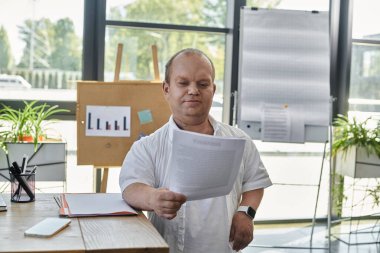 Beyaz gömlekli bir adam masanın başında duruyor, modern bir ofiste belgeleri dikkatle inceliyor..