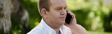 Beyaz gömlekli bir adam yeşil bir bahçede dikiliyor, dikkatle bir telefon konuşmasını dinliyor..