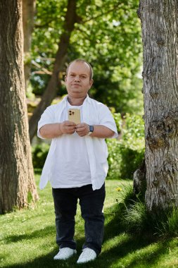 Çevresi yeşil ağaçlar ve otlarla çevrili bir parkta akıllı telefon tutan bir adam..