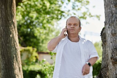 Beyaz tişörtlü bir adam parkta yürüyor, telefonuyla konuşuyor..