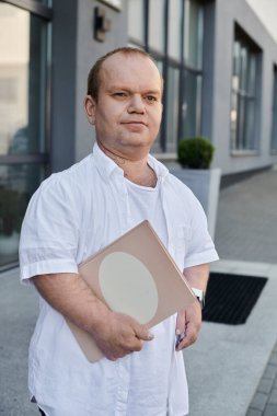 Beyaz gömlekli bir adam elinde klasörle modern bir binanın önünden geçiyor..